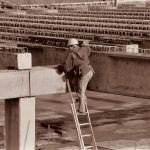 Trabalhador usando equipamentos de segurança sobe uma escada vertical em uma estrutura industrial.