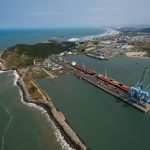 Vista aérea do porto de Imbituba, com cais, guindastes, navios e a faixa costeira ao redor.