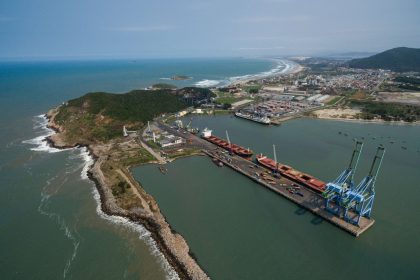 Vista aérea do porto de Imbituba, com cais, guindastes, navios e a faixa costeira ao redor.