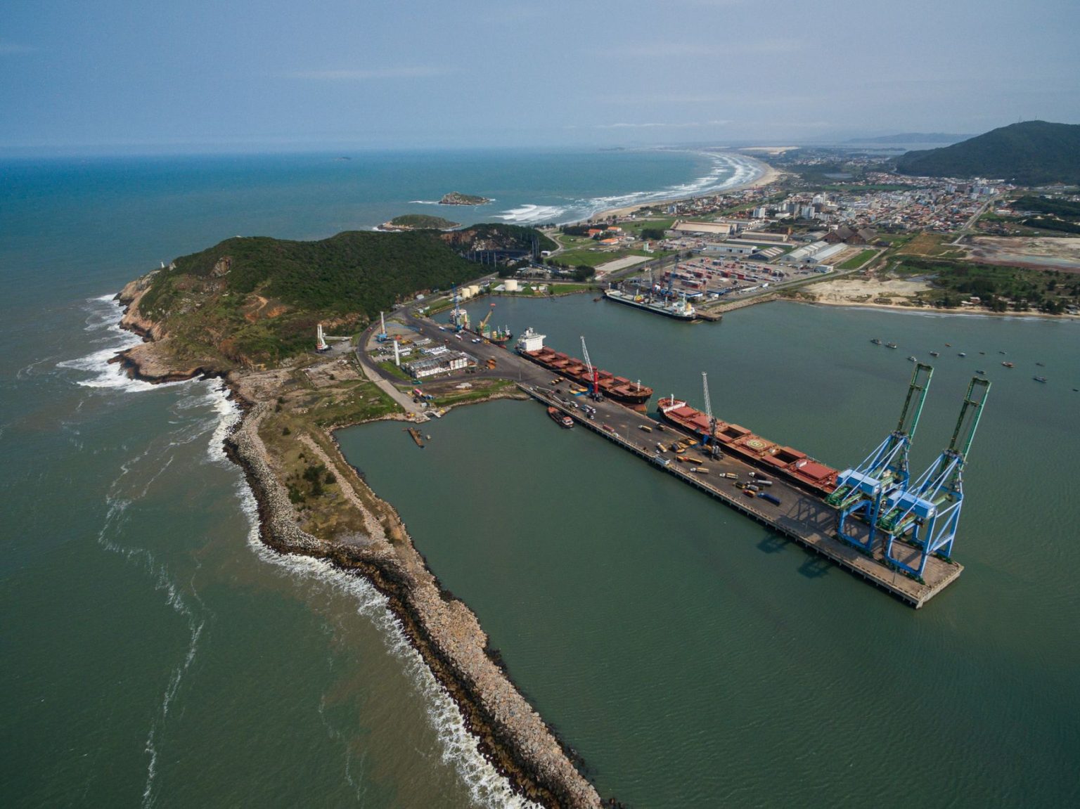 Vista aérea do Porto de Imbituba, em Santa Catarina, com navios, guindastes, instalações portuárias e a faixa litorânea.